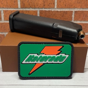 May include: A black firearm magazine with a single bullet visible, resting on a brown surface. Below it is a green patch with the text "Haterade" in white and orange, resembling a sports drink logo.