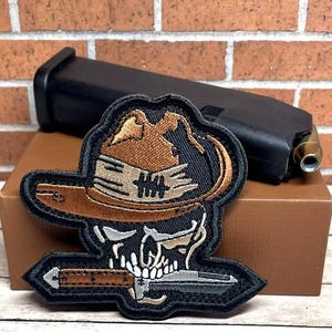May include: A black and brown embroidered patch featuring a skull wearing a brown cowboy hat and holding a cigar. The skull has a bandana around its neck and a mustache.