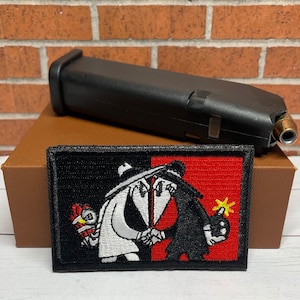 May include: A black and white embroidered patch featuring two figures, one in a white suit and the other in a black suit, holding a red and yellow bomb. The patch is on a red background with a yellow star.