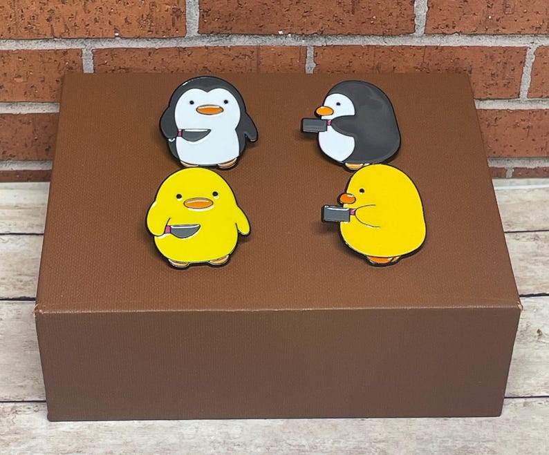 May include: Four enamel pins featuring cartoon animals holding a knife. The pins are on a brown surface. The animals are a penguin, a chick, and two penguins.