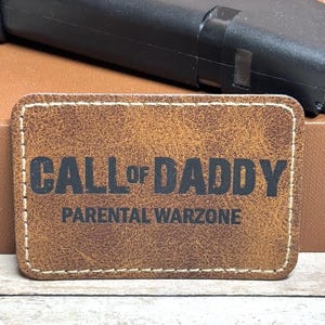 May include: A rectangular brown leather patch with the text "CALL OF DADDY" above "PARENTAL WARZONE" in black. The patch has a stitched border and a textured surface. A black object is in the background.