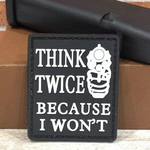 May include: Black square patch with white text that reads "THINK TWICE BECAUSE I WON'T." A white graphic of a handgun is on the right side. A black object is in the background.