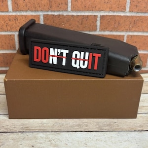 May include: A black magazine and a rectangular patch with the words "DON'T QUIT" in white and red lettering. The patch is on a brown box, with a brick wall background.