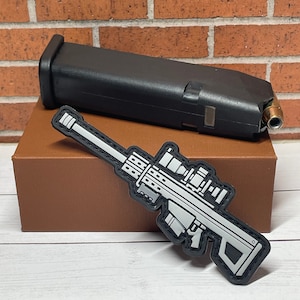 May include: A white and gray sniper rifle patch with a black background. The patch is on a brown box.