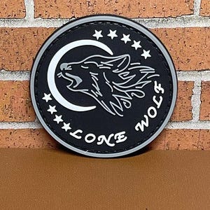 May include: Black and white embroidered patch with a wolf howling at the moon. The patch has five stars around the moon and the words "Lone Wolf" written below the moon.