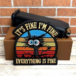Peut inclure: Patch brod&eacute; noir avec un personnage de dessin anim&eacute; d&eacute;labr&eacute; aux grands yeux et le texte "It's Fine I'm Fine Everything Is Fine" dans un design de coucher de soleil r&eacute;tro.