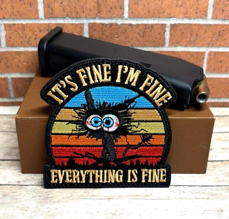 Peut inclure: Patch brod&eacute; noir avec un personnage de dessin anim&eacute; d&eacute;labr&eacute; aux grands yeux et le texte "It's Fine I'm Fine Everything Is Fine" dans un design de coucher de soleil r&eacute;tro.