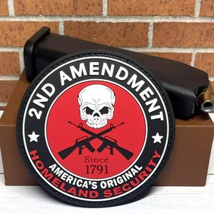 May include: Black and red round patch with the text "2nd Amendment" in white. The patch features a skull and crossbones with two rifles, and the text "Since 1791 America's Original Homeland Security".