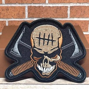 May include: Embroidered patch featuring a skull with crossed axes and a comb design. The skull is tan with black markings, and the axes are silver. The patch has a black border and is likely for jackets or bags.