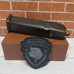 May include: A black plastic magazine for a handgun, sitting on a brown box with a black patch that says "Bulldog Tactical Gear" with a bulldog graphic and the text "Fac Fortia Et Patere".