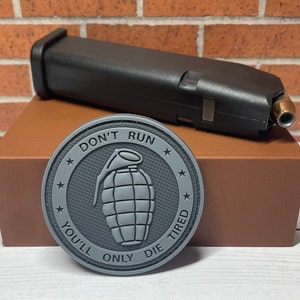 May include: A gray, circular patch with the text "DON'T RUN YOU'LL ONLY DIE TIRED" and a grenade graphic. A black magazine with a bullet is resting on a brown box. The background is a brick wall.