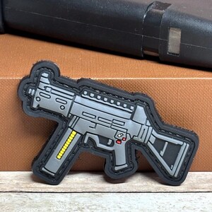 May include: A gray and black patch in the shape of a submachine gun. The patch has a black border and features details like the magazine, trigger, and sights. The magazine has yellow accents. The background includes a black object.