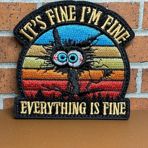 Peut inclure: Patch brod&eacute; avec un personnage de dessin anim&eacute; noir et blanc aux grands yeux et un air de d&eacute;tresse. Le personnage est devant un fond de couleur coucher de soleil avec le texte "It's Fine I'm Fine Everything Is Fine" en fil dor&eacute;.
