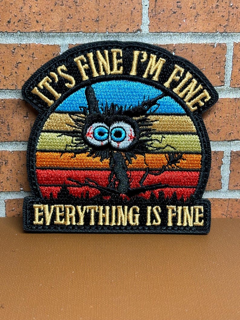 Peut inclure: Patch brod&eacute; avec un personnage de dessin anim&eacute; noir et blanc aux grands yeux et un air de d&eacute;tresse. Le personnage est devant un fond de couleur coucher de soleil avec le texte "It's Fine I'm Fine Everything Is Fine" en fil dor&eacute;.