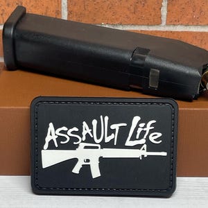 May include: Black patch with white text that reads "Assault Life" and a white silhouette of a rifle.