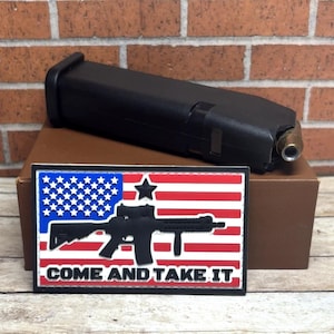 May include: Black firearm magazine with a brass-colored tip, resting on a brown surface. Below, a patch featuring the American flag, a rifle silhouette, and the text "COME AND TAKE IT" in white.
