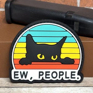 May include: A semi-circular rubber patch featuring a black cat silhouette with yellow eyes peeking over a horizontal striped background of teal, yellow, and orange. Below the cat, the text "EW, PEOPLE." is printed in black on a white background.