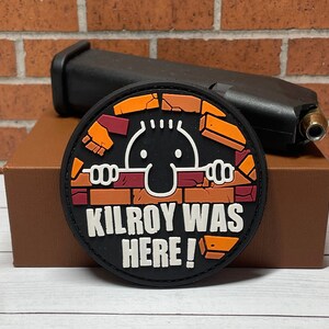 May include: A black and orange round patch with a cartoon character peeking over a brick wall. The text "Kilroy Was Here!" is printed on the patch.