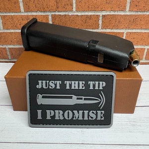 May include: A black and gray patch with the text "Just the tip I promise" and a gray silhouette of a bullet. The patch is on top of a brown box with a black magazine on top of the box.