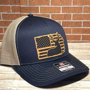 May include: A navy blue and tan trucker hat with a brown leather patch featuring an American flag with a deer silhouette. The hat is embroidered with the text "Richardson 112 Adjustable".
