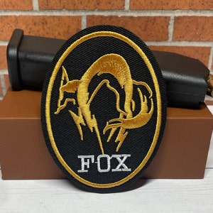 May include: A black and gold embroidered patch with a stylized fox design and the word "FOX" in white lettering.