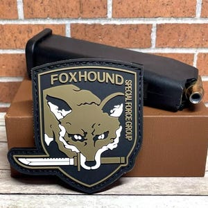 May include: A black and tan embroidered patch with a fox head and the text "FOXHOUND SPECIAL FORCE GROUP".