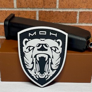 May include: Black and white embroidered patch with a bear head and the letters "MOH" in a shield shape.