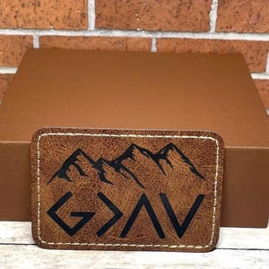 May include: A brown leather patch with a stitched border. The patch features a mountain range design with the word "GOD" in black lettering and an arrow pointing upwards.