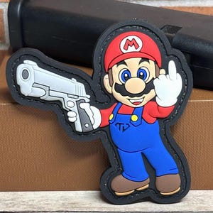 May include: A black-bordered patch featuring a cartoon character in a red hat and blue overalls, holding a silver handgun and giving the middle finger. The character has a mustache and is smiling. A black object is in the background.