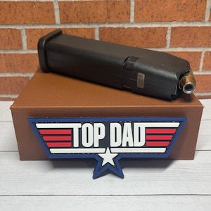 May include: A black firearm magazine with a single bullet visible, resting on a brown box. A patch with the words "TOP DAD" in white, red, and blue is below the box, with a blue star at the bottom.