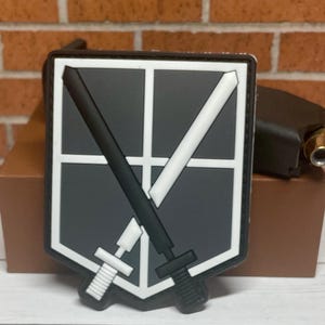 May include: A black and white patch with a shield design and two crossed swords. The shield has a gray background and white lines. The swords are black with white handles.