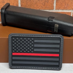 May include: A black and gray American flag patch with a red stripe across the middle. The patch is on a brown surface.