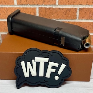 May include: A black plastic magazine for a handgun with a brass bullet casing on top. The magazine is sitting on a brown box with a black patch that says "WTF!" in white letters.