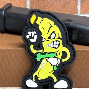 May include: A yellow and black patch featuring an angry banana character with white teeth and a green accessory. The patch has white gloves and shoes. The text "5.11" is visible on the green accessory.