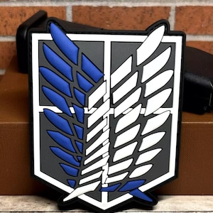 May include: A black, white, and blue rubber patch with a winged emblem. The wings are stylized and have a gray background. The patch is shaped like a shield.