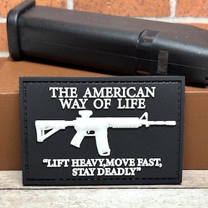 May include: Black rectangular patch with white text and a white rifle graphic. The text reads "THE AMERICAN WAY OF LIFE" and "LIFT HEAVY, MOVE FAST, STAY DEADLY". A black object is in the background.
