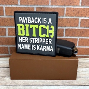 May include: A black and gray patch with the text "PAYBACK IS A BITCH HER STRIPPER NAME IS KARMA" in white and neon green lettering. The patch is on a brown box, with a black object to the right.