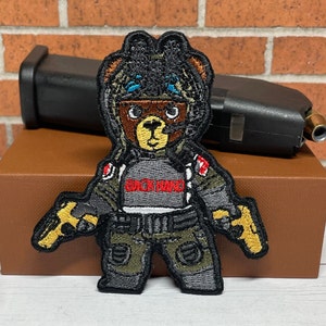 May include: A brown and black embroidered patch of a bear wearing a helmet and holding two guns. The bear is wearing a grey and green tactical suit with the text "BACK HAND" on the chest.