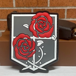 May include: A gray and white patch with a black border, featuring two red roses with white stems and leaves. The patch has a gray background with a white outline and a black border.