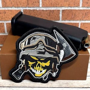 May include: Black and white embroidered patch of a skull wearing a helmet with a pickaxe. The skull has yellow teeth and eyes.