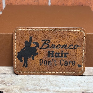 May include: Brown leather patch with a black silhouette of a cowboy riding a bucking bronco. The text "Bronco Hair Don't Care" is written in black lettering.