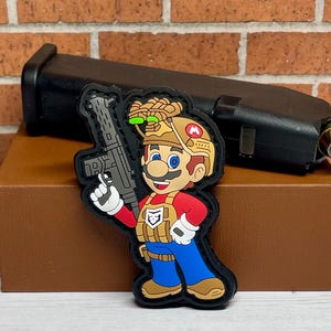 May include: A black and brown tactical patch featuring a cartoon character wearing a helmet and holding a gun. The character has a red shirt, blue pants, and a brown vest. The patch is made of a durable material and is perfect for adding a unique touch to your gear.