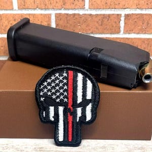 May include: Black firearm magazine with a gold-colored bullet at the end, resting on a brown box. A black, white, and red embroidered patch in the shape of a skull with a flag design is in front of the magazine.