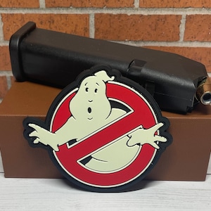 May include: A red and white Ghostbusters logo patch with a cartoon ghost in the center. The patch is attached to a brown leather box.