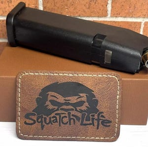 May include: A brown leather patch with a black silhouette of a Bigfoot and the text "Squatch Life". The patch is on a brown box with a black magazine on top.
