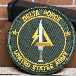 May include: A circular Delta Force United States Army patch. The patch features a green background with gold embroidered text and accents. The central design includes a white blade, a yellow lightning bolt, and a yellow triangle.