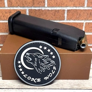 May include: A black and gray patch with a wolf howling at the moon and the text "Lone Wolf" around the edge. The patch is on a brown box with a black magazine for a gun on top of it.