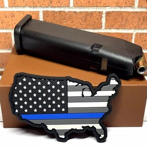 May include: A black and white American flag patch with a blue line across the bottom, shaped like the United States. The patch is on a brown surface next to a black magazine.