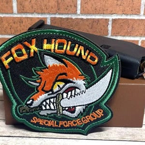 May include: Green, black, and orange embroidered patch with the text "FOX HOUND SPECIAL FORCE GROUP" and a cartoon fox with a knife in its mouth.