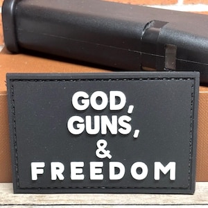 May include: Black rectangular patch with the words "GOD, GUNS, & FREEDOM" in white, raised lettering. A black object is in the background. The patch is likely a morale patch or a decorative item.